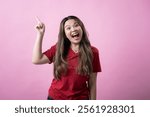 Asian woman in a red t-shirt and jeans standing against a pink background, smiling brightly with excitement, raising one finger upward as if having a new idea or solution