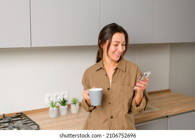 Asian Woman Relaxing In The Kitchen With Cup Of Coffee, Checking Messages On The Smartphone, Scrolling News Feed Drinking Morning Coffee. Smiling Girl Texting Online