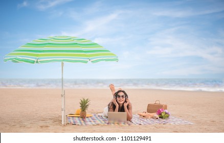 Asian Woman Relaxing And Listening To Music  Headphone Form Laptop Smartphone, Lying On Beach Mat In The Beach.