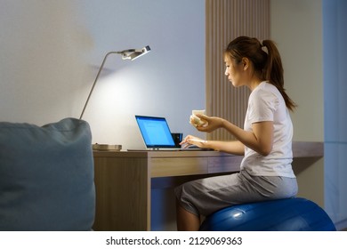 Asian Woman Relaxing On Yoga Ball And Eating Sandwich And Chatting Online With Team Of Colleagues On Laptop While Work From Home At Home At Night.