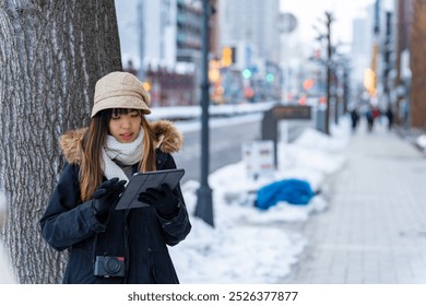 Asian woman remote working on digital tablet during waiting for bus in winter night. Attractive girl enjoy urban outdoor lifestyle travel in the city with using wireless technology and gadget device. - Powered by Shutterstock