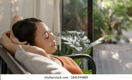 Asian woman resting breathing fresh air feeling mental balance enjoying wellbeing at home on sofa, closed eyes put hands behind head relaxing on comfortable sofa in cozy warm light living room