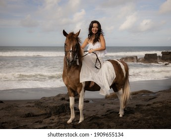 Asian woman riding horse on the beach. Outdoor activities. Woman wearing long white dress. Traveling concept. Cloudy sky. Copy space. Bali, Indonesia