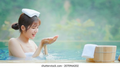 asian woman scoop up the water and feel the drop while sitting in hot spring with towel on head