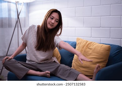 Asian woman seated on a blue sofa stretch her legs reaching for her toes recovery hamstring in a cozy softly lit living room at night before sleep - Powered by Shutterstock