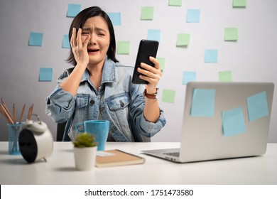 Asian Woman Shocked Looking At Smart Phone Reading News And Screaming At Home Office. WFH. Work From Home. Prevention Coronavirus COVID-19 Concept.