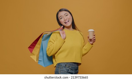 Asian woman showing shopping bags on camera, holding cup of coffee in hand. Shopaholic adult carrying paper bags after making purchase on discount sale at retail store. Cheerful customer