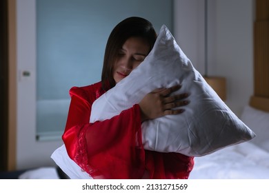 Asian Woman In A Silk Red Nightgown Standing Hugging Pillow With Two Hands In A Bedroom At Night. Female In Satin Nightwear And Embracing Soft Pillow