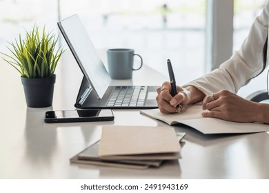 An Asian woman sits at her office desk and uses a pen to write down appointments in her notebook as a reminder. Notebooks are still popular for note taking and are used a lot in school. - Powered by Shutterstock