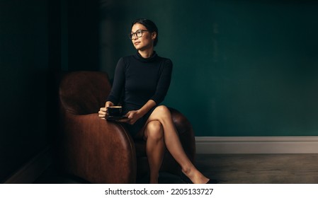 Asian woman sitting in armchair with cup of coffee and looking away. Thoughtful businesswoman relaxing in her office. - Powered by Shutterstock