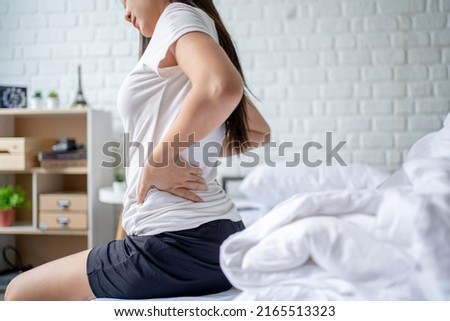 Asian woman sitting in bed touching her back, feeling back pain in the morning. She suffered from pain in the lumbar muscles after waking up.
