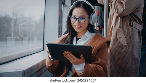 Asian woman sitting next to large window of moving train. Wearing headphones for music while using tablet device. Playing game or browsing web. Enjoy during trip on public transportation. - Powered by Shutterstock