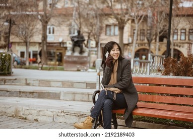 Asian Woman Sitting On A Bench In The Park While Using Her Mobile Phone