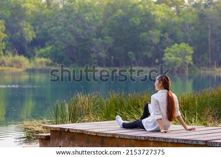 Asian woman sitting on the edge of dock with peaceful natural park during summer looking at turquoise lake for serene and relaxation outdoor recreation concept