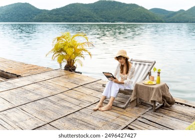 Asian woman sitting on lake house balcony and using digital tablet remote working online business or watching movie. Attractive girl enjoy outdoor lifestyle travel nature on summer holiday vacation. - Powered by Shutterstock