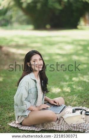 Asian woman sitting on picnic blanket and lawn at park working on laptop. Asian female using laptop while sitting under a tree at park with bright sunlight. Work from anywhere concept.