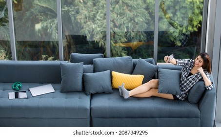 Asian woman sitting on sofa near big glass windows, relaxing alone in house with green forest in background, taken from high angle.