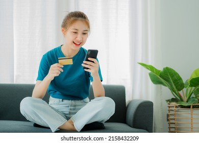 Asian Woman Sitting On Sofa Holding Credit Card And Shopping From Smartphone Online. At Home For Online Banking Use Mobile Online Shopping