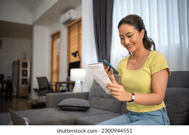 Asian woman sitting on sofa with mobile phone paying bills at home
