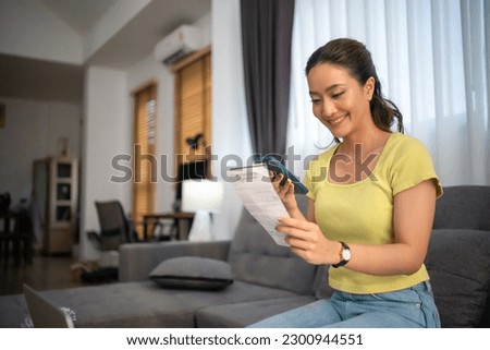 Asian woman sitting on sofa with mobile phone paying bills at home