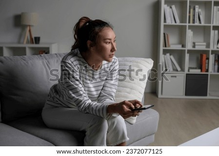 Asian woman sitting on sofa holding phone feeling disappointed, sad, upset. Female suffer from mental health problems.