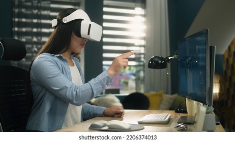 Asian Woman Sitting At The Table At Home Office And Using VR Headset To Work On 3D Modeling Project On Personal Computer. Virtual Reality. Metaverse