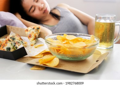 Asian woman sleep after eating junk food with pizza, potato chips and glass of beer on desk, bad habit, unhealthy lifestyle concept 