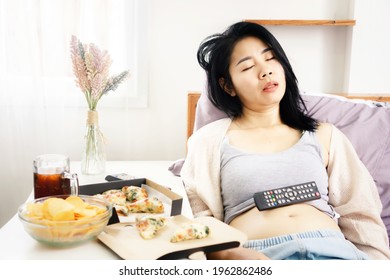 Asian Woman Sleeping In Bed After Eating Pizza, Potato Chips And Soda With Tv Remote On Her Fat Belly  Overeating Junk Food Concept 