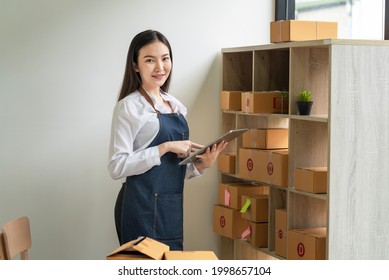 Asian Woman Small Business Owner Holding A Tablet To Check Parcels Before Delivering To Customers At Home. Looking At Camera.