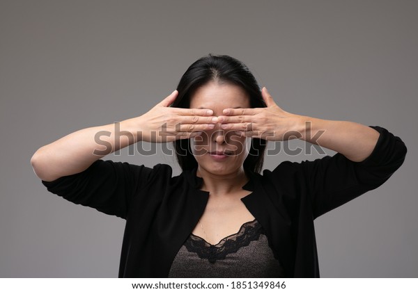 Asian woman in a smart\
black outfit covering her yes with her hands in a concept of the\
metaphor see no evil, hear no evil speak no evil, over a grey\
studio background