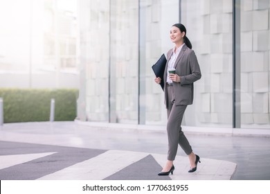 Asian Woman With Smartphone Standing Against Street Blurred Building Background. Fashion Business Photo Of Beautiful Girl In Casual Suite With Phone And Cup Of Coffee