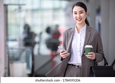 Asian Woman With Smartphone Standing Against Street Blurred Building Background. Fashion Business Photo Of Beautiful Girl In Casual Suite With Phone And Cup Of Coffee