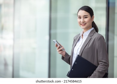 Asian Woman With Smartphone Standing Against Street Blurred Building Background, Asian Business Photo Of Beautiful Girl In Casual Suite With Smart Phone. 