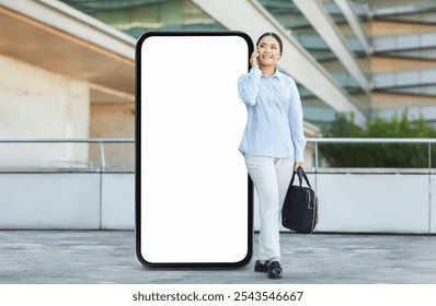 Asian woman smiles while talking on a phone, standing next to a large blank phone screen in an outdoor urban setting, dressed in casual attire, ready for her day ahead.