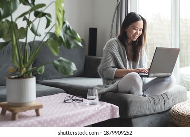 Asian woman smiles while working from home on her laptop computer, sitting on a sofa in the living room of a house in Edinburgh, Scotland, United Kingdom - Shutterstock ID 1879909252