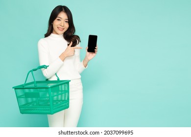 Asian woman smiling and holding grocery basket and presenting mobile phone application isolated on green background, Shopping and Supermarket concept