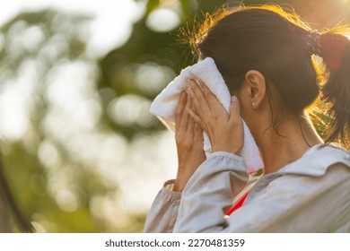 Asian woman in sportswear wiping sweat on her face with towel during jogging exercise at public park at summer sunset. Healthy girl enjoy outdoor lifestyle sport training workout running in the city.