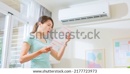 asian woman stand by air conditioner shocked and upset about electricity bill in hand at home - economic inflation concept