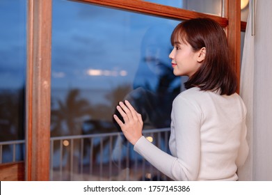 Asian Woman Standing In Apartment Home. Thai Female Looking Through Window To See Seaview On Sunset. Wellness And Relax In Bedroom.