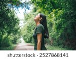 An Asian woman is standing happily in a green forest park, enjoying the fresh air and nature. She takes a deep breath, looks up at the morning sunlight, and feels relaxed and peaceful.