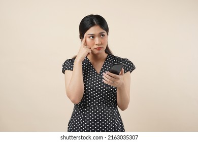 An Asian Woman Standing Infront Of Cream Background, Holding Her Smartphone With A Confuse Expression. Isolated On Cream Background. Close Up. 