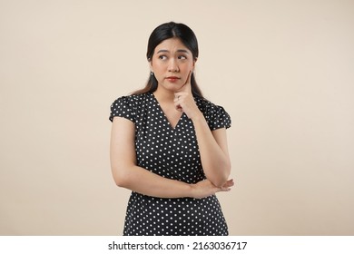 An Asian Woman Standing Infront Of Cream Background, With A Thinking Gesture And Rolling Eyes. Isolated On Cream Background. Close Up. 