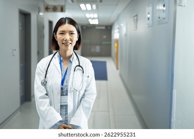 Mujer asiática de pie y sonriente en el pasillo del hospital, con bata de laboratorio, estetoscopio e insignia de identificación. Cuidado de la salud, médico, clínico, profesionalismo, confianza, tranquilidad, bienvenida