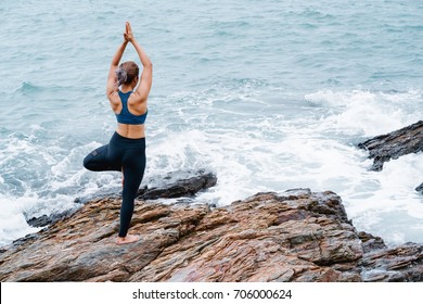 Asian Woman Standing Yoga Postures Tree (vrks Asana) On The Rocks. Sea And Sky As Background.