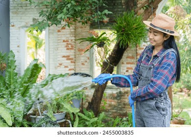 Asian woman, straw hat, waters lush greenery with hose. Satisfied smile, blue gloves on, tranquil ambiance amidst verdant plants and brick wall. flower garden owner Taking care of trees for sale - Powered by Shutterstock