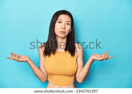 Asian woman in summer yellow top, studio setup, doubting and shrugging shoulders in questioning gesture.