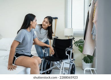 Asian woman support female leg amputee friend sitting on wheelchair. Attractive happy young girl legless patient doing physiotherapy indoors with roommate in bedroom at home. Leisure activity in house - Powered by Shutterstock