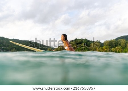 Asian woman surfer paddling surfboard and riding the wave in the sea at tropical beach at sunset. Healthy female enjoy outdoor activity lifestyle and water sport exercise surfing on summer vacation