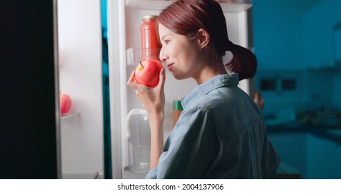 asian woman takes apple from opened refrigerator for late night supper in kitchen at night