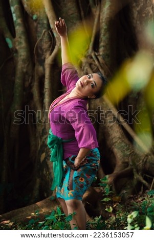 Asian woman takes a dancing pose in the jungle while wearing a purple dress and a golden necklace with a beautiful smile on her face